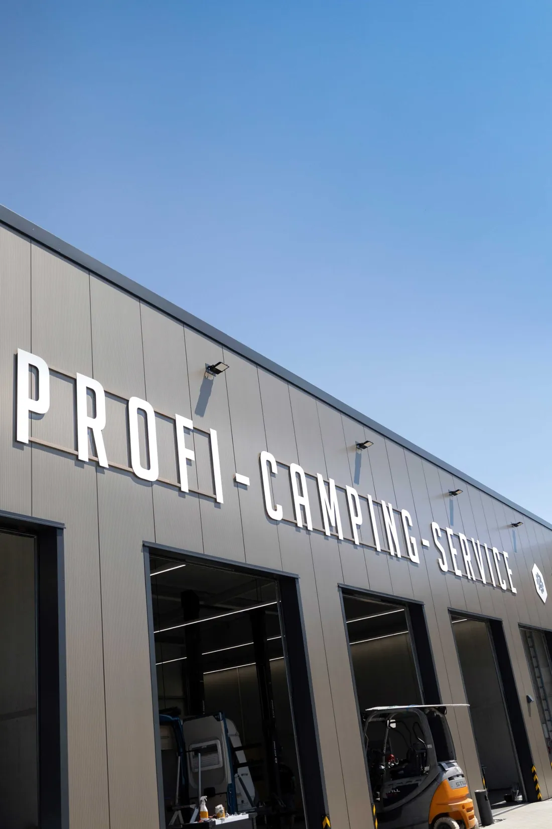 Fassade des Profi-Camping-Service mit weißem Schriftzug und geöffneten Hallentoren, im rechten Tor steht ein Gabelstapler
